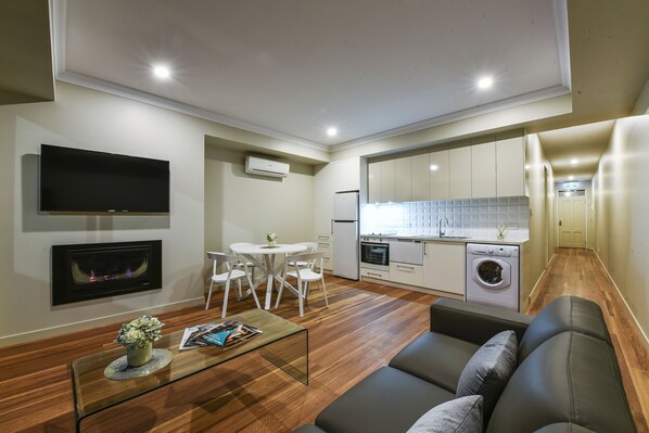Un espacio habitable compacto con sofá, una mesita de centro de vidrio, un televisor de pantalla plana, chimenea, zona de comedor con mesa y sillas blancas, y kitchenette con refrigerador, horno y lavadora.