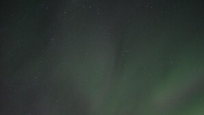 Une cabane dotée d’une porte et de fenêtres bien visibles, entourée d’arbres, sous un ciel nocturne où l’on peut apercevoir des étoiles et des aurores boréales.