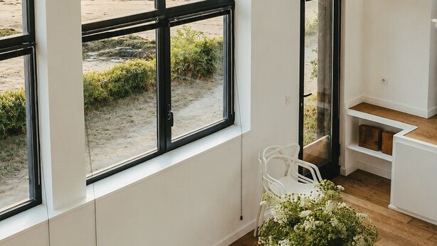 A room with a large window overlooking a body of water, a glass table with chairs, and a white chair with a vase of flowers. - A boat house on the beach (Paimpol)