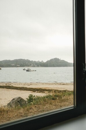 A view of a beach with a sandy shore and a body of water. There are boats visible on the water, and a hill with trees in the background. - A boat house on the beach (Paimpol)