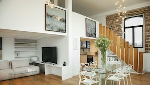 A modern living room with a glass coffee table, white sofa, and a TV. There's a staircase with wooden panels and a large window. - A boat house on the beach (Paimpol)