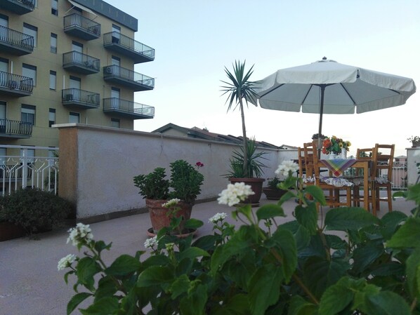 Een terras met een tafel en stoelen onder een grote parasol, omringd door potplanten en bloemen.