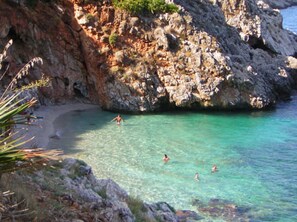 A rocky coastline with clear turquoise water, a sandy beach, and people swimming. - Holiday house with garden 1 km. from the beach. (Castellammare del Golfo)