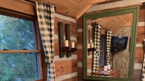 L’intérieur d’une cabane, avec une commode en bois, une chaise, un miroir et une fenêtre ornée de rideaux à carreaux.