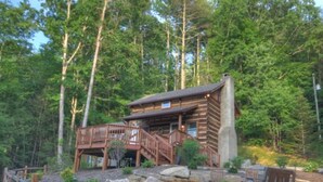 Une cabane en rondins avec une terrasse en bois et des fondations en pierre, entourée d’une forêt dense.