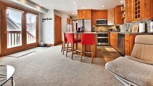 Eine moderne Küche mit Holzausstattung, einem Edelstahlkühlschrank und einer Mikrowelle. Im Wohnbereich befinden sich zwei rote Barhocker und ein beiges Sofa.