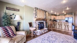 A cozy living room with a fireplace, a dining area with wooden chairs, and a kitchen in the background.