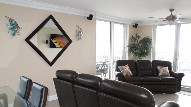 A living room with a glass dining table, two brown leather sofas, a ceiling fan, and large windows. - Completely Remodeled Corner Unit With Amazing Gulf Views! (Destin)