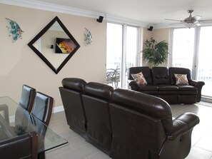 A living room with a glass dining table, two brown leather sofas, a ceiling fan, and large windows.