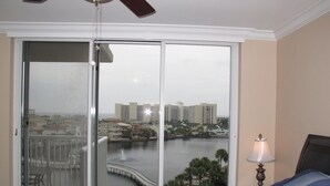 A bedroom with a large sliding glass door leading to a balcony, a bed with blue bedding, a nightstand with a lamp, and a view of a river and buildings. - Completely Remodeled Corner Unit With Amazing Gulf Views! (Destin)