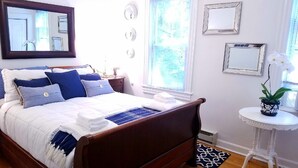 A neatly made bed with blue and white bedding, a wooden headboard, and a small white table with a potted plant beside the window. - Great South End / Back Bay location, 1 bed condo near T station  (Boston)