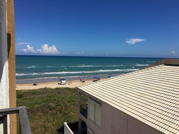 A beach view from a building with a tiled roof, a grassy area, and a clear blue sky.