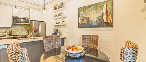 A modern kitchen with white cabinets, a microwave, and a refrigerator. There is a dining table with wicker chairs and a bowl of fruit. A painting of sailboats hangs on the wall.