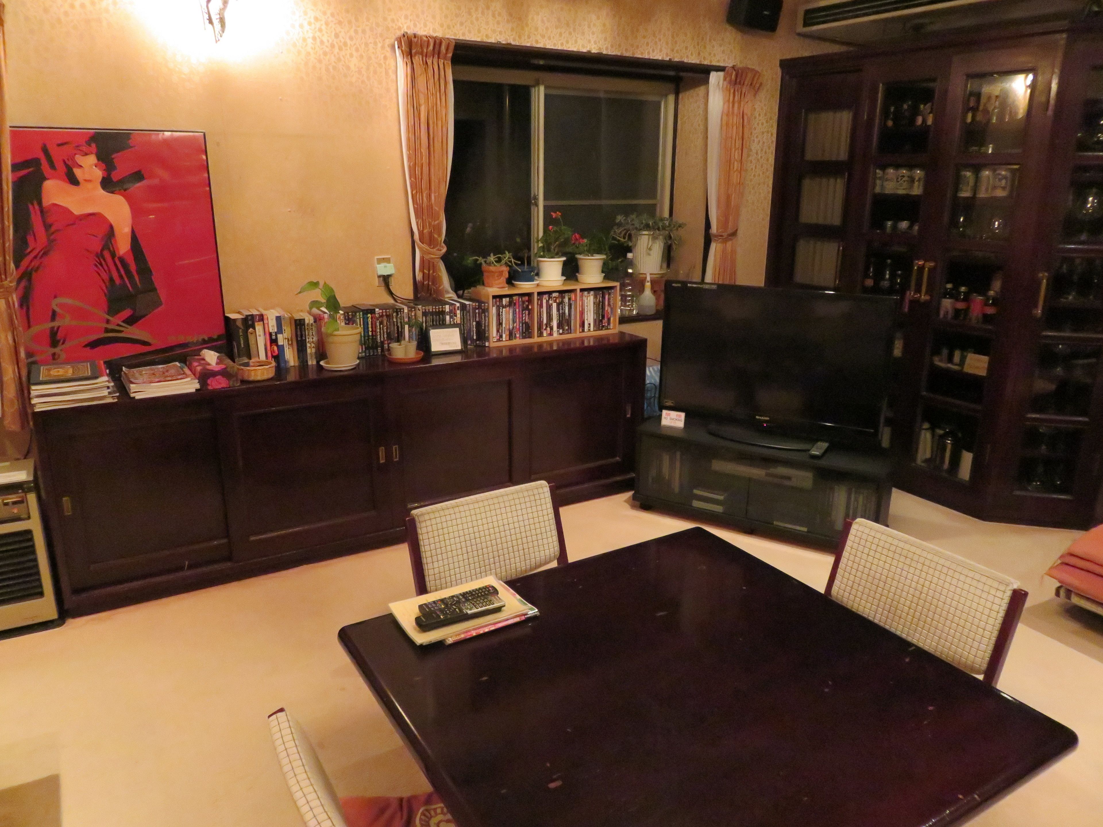 Un salón con una mesa de madera oscura, dos sillas de malla metálica, un televisor de pantalla plana, una estantería y una ventana con cortinas.
