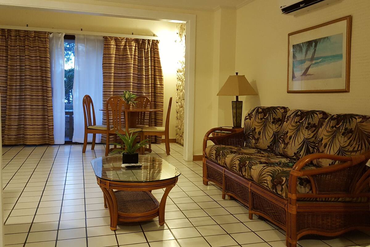 A living room with a glass-top dining table, wicker furniture, a sofa with a floral pattern, and a painting on the wall.