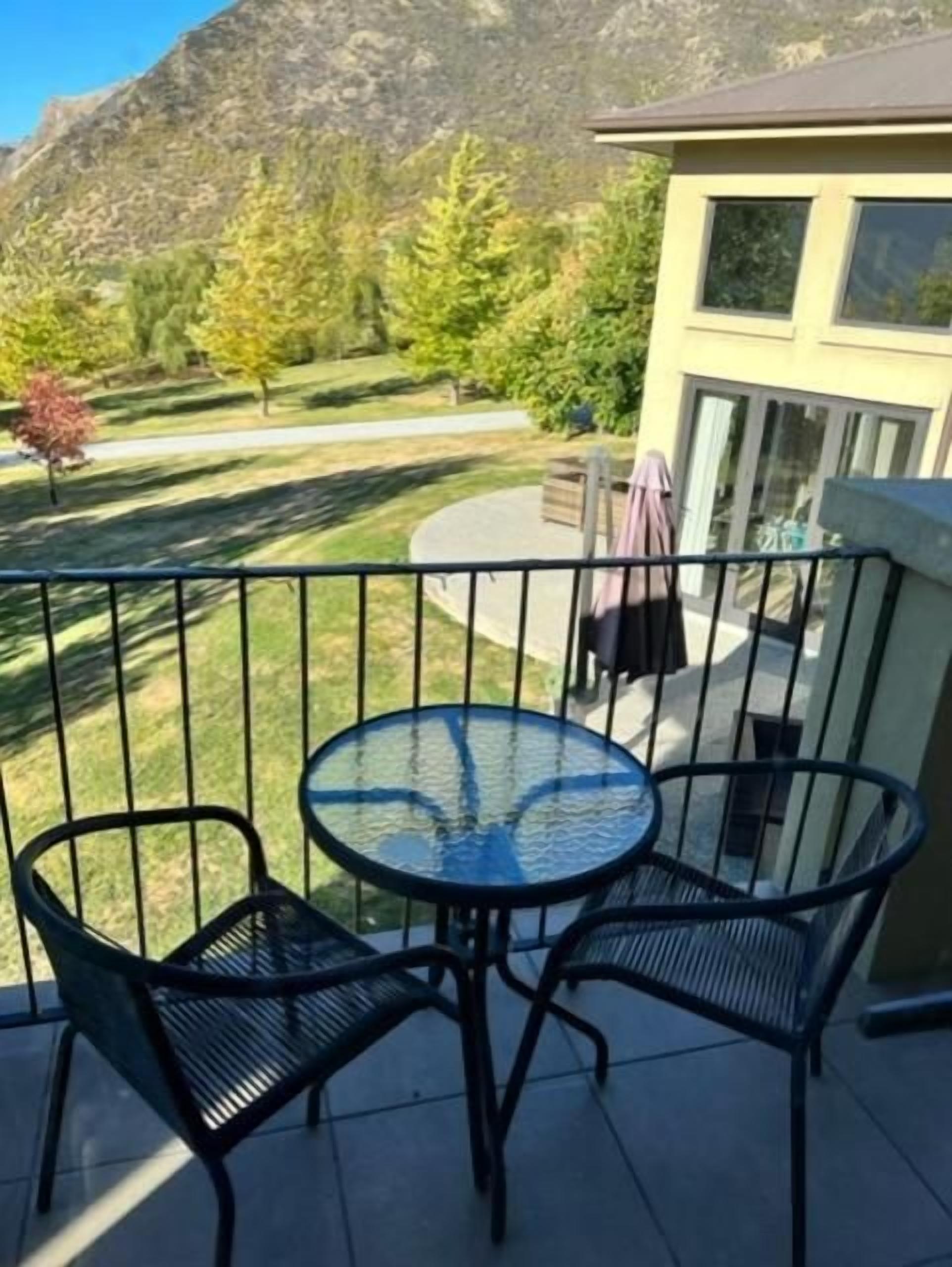 A balcony with a glass table and chairs, overlooking a mountain and a well-maintained lawn.
