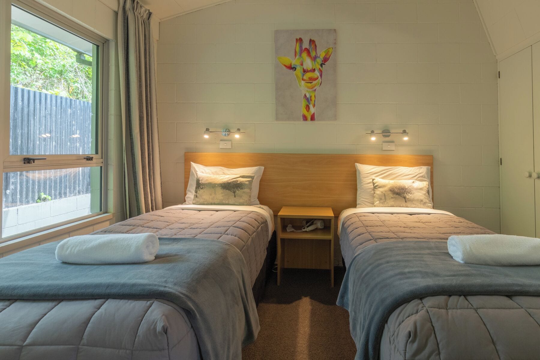 A bedroom with two beds, a wooden headboard, bedside tables, and a painting of a giraffe.