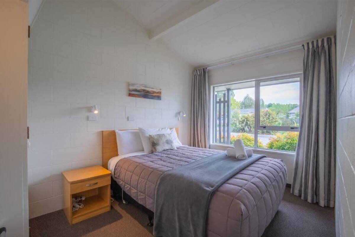 A bedroom with a bed, a nightstand, a window with curtains, and a painting on the wall.