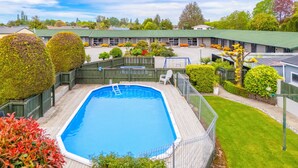 A swimming pool with a wooden deck, surrounded by a fenced area with a lawn and various shrubs.