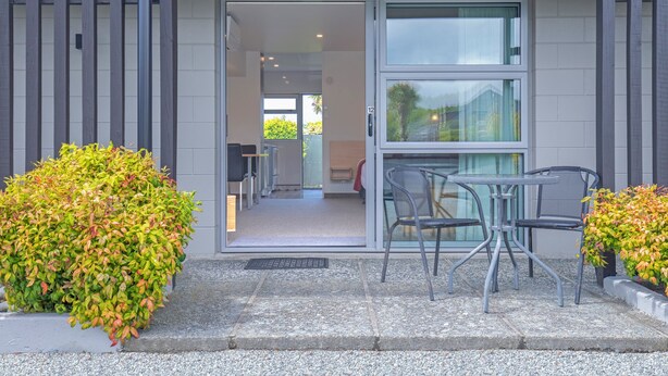 A modern building entrance with a glass door, a small outdoor seating area with metal chairs, and a gravel pathway.