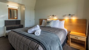 A modern hotel room with a large bed, a wooden headboard, a nightstand, a mirror, and a bench with a towel on it.