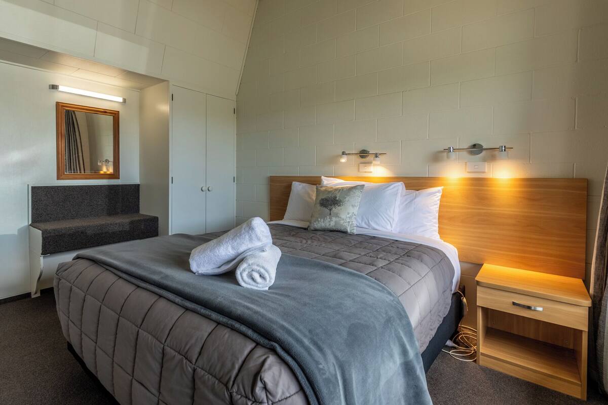A modern hotel room with a large bed, a wooden headboard, a nightstand, a mirror, and a bench with a towel on it.