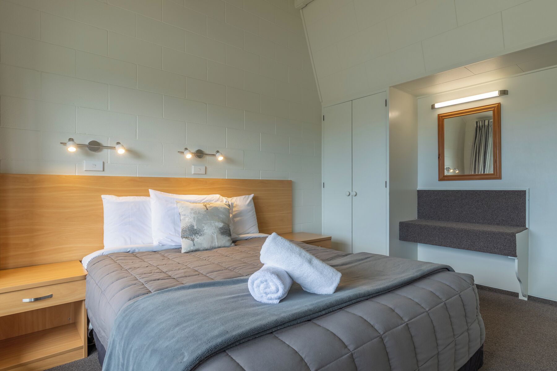 A neatly arranged hotel room with a large bed, a bedside table, a nightstand, a mirror, and a built-in bench.