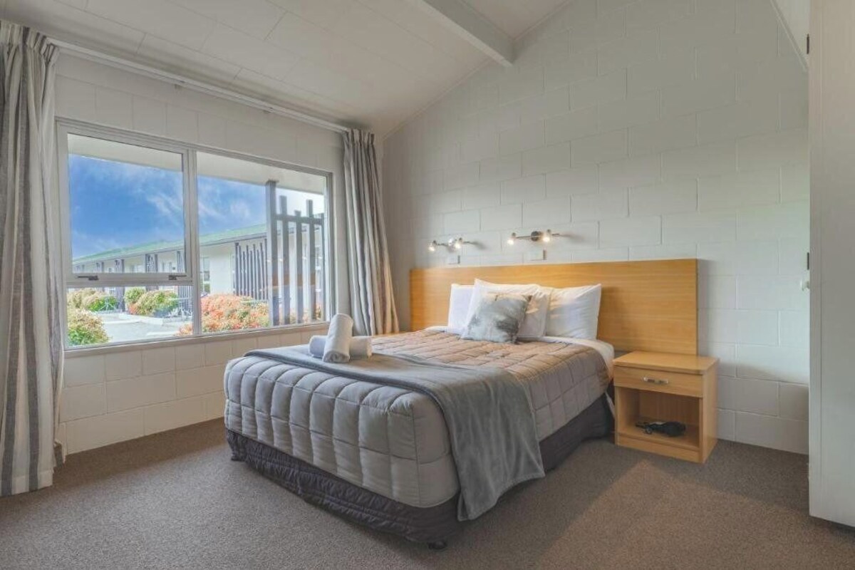 A bedroom with a bed, a nightstand, a window with curtains, and a brick-patterned wall.