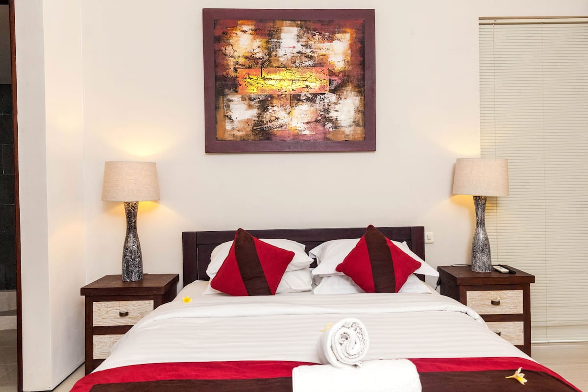 Un lit bien fait, avec une literie rouge et blanche, deux lampes de chevet, un tableau au mur et une fenêtre fermée.