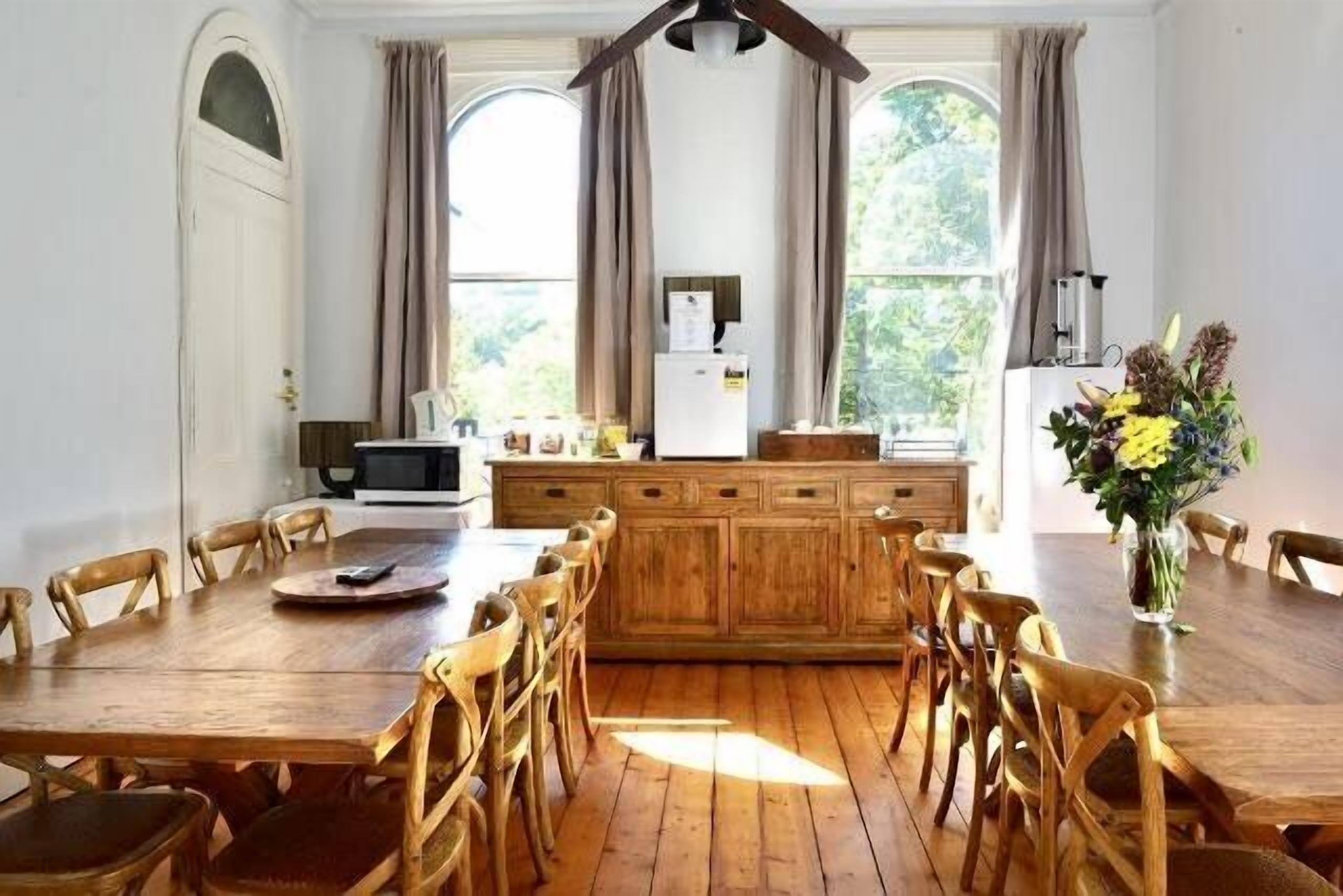 A dining area with a wooden table and chairs, a kitchen in the background, and a large window with curtains.