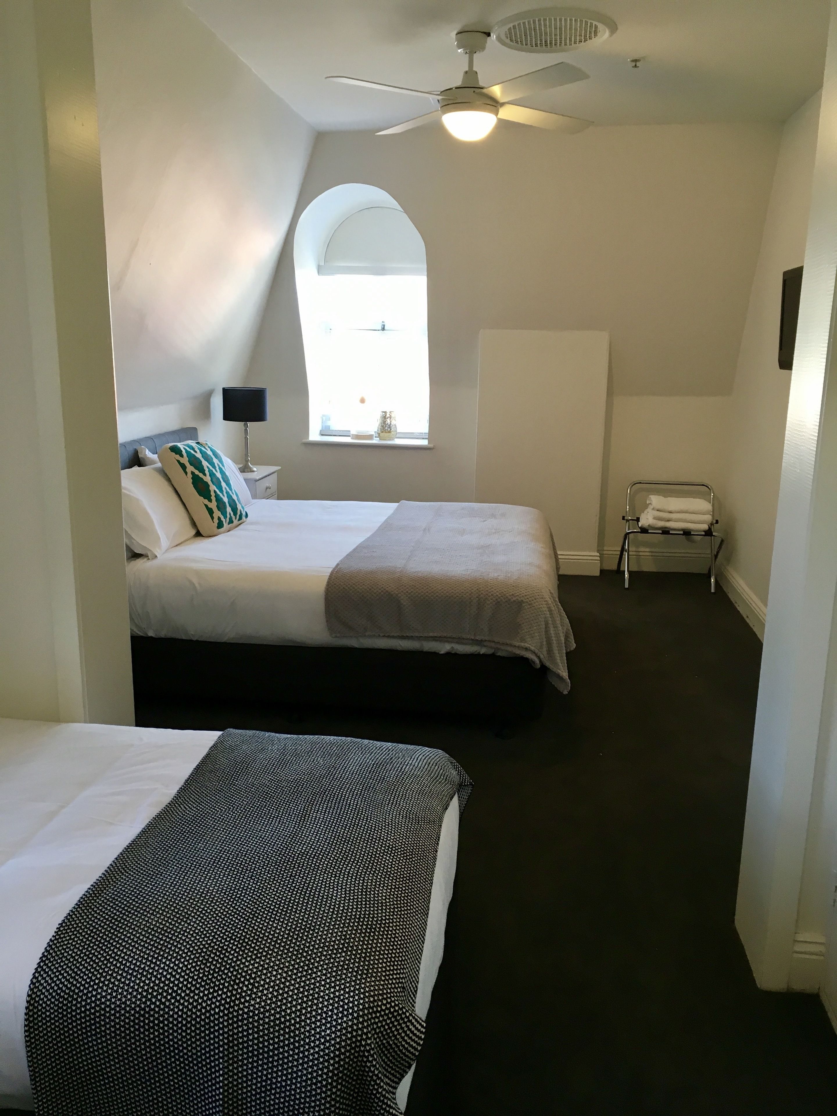 A hotel room with two beds, a ceiling fan, a window with natural light, a small table with a lamp, and a towel rack.