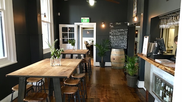 Un interior de restaurante con una larga mesa de madera, varios taburetes, un menú en una pizarra y una vitrina con vino.