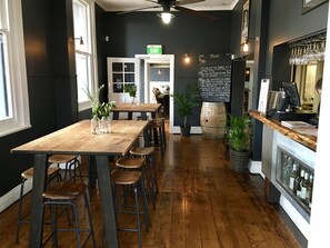 A restaurant interior with a long wooden table, multiple stools, a chalkboard menu, and a wine display.