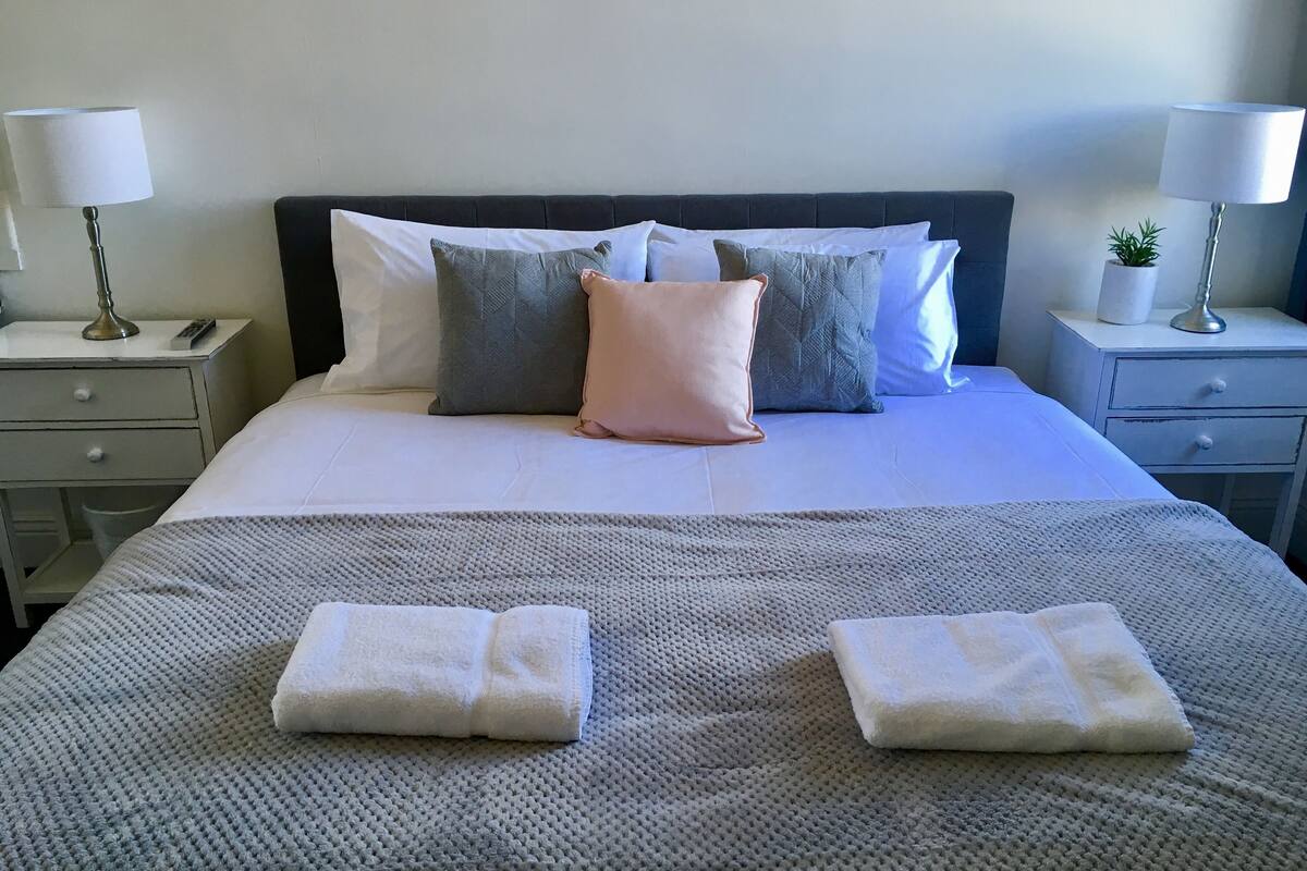 Un lit bien fait, recouvert d’une couverture grise, avec deux oreillers et deux serviettes pliées. Il y a deux tables de chevet, chacune avec une lampe, et une petite plante sur l’une d’elles.