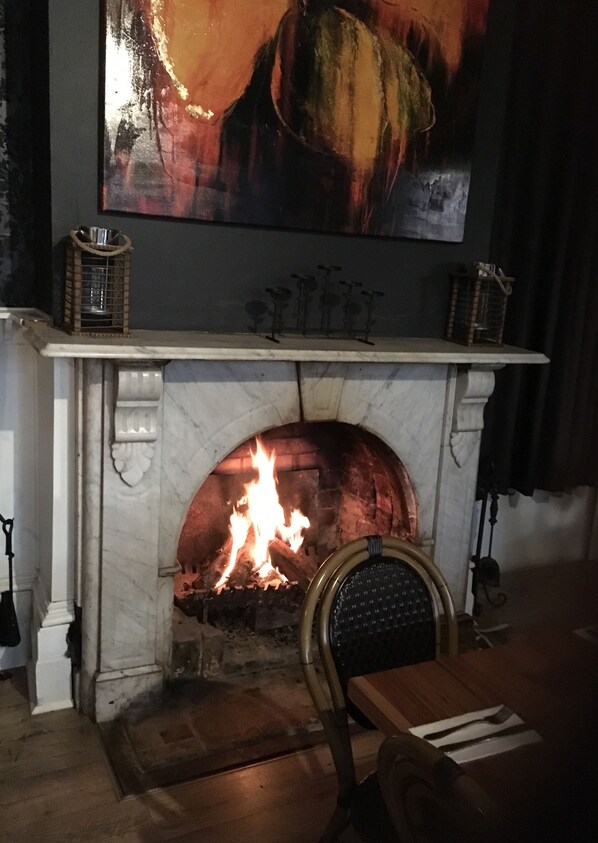 A fireplace with a fire, a table, and chairs in a room with a dark wall and a painting above the fireplace.