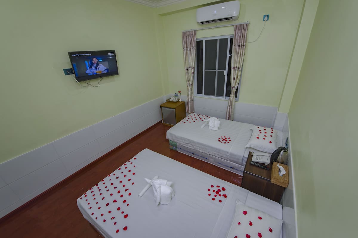 Une chambre d’hôtel avec deux lits, une télévision, une fenêtre avec des rideaux, une table de chevet avec un téléphone et un thermos, et un climatiseur fixé au mur.