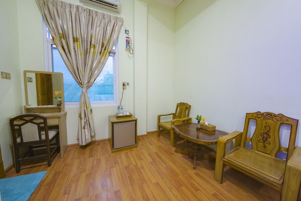 Una habitación con piso de madera, un escritorio con espejo, un refrigerador pequeño, una mesa de comedor con sillas y una ventana con cortinas.