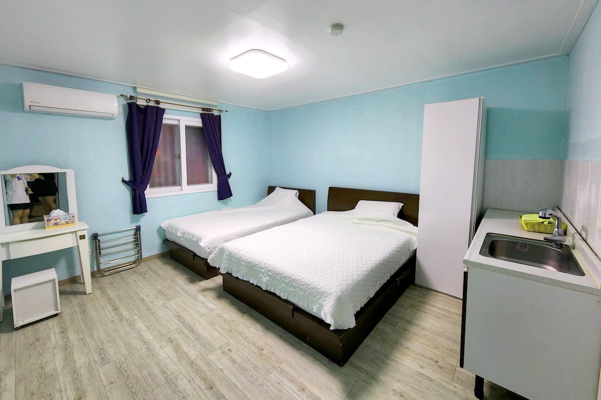 A bedroom with a double bed, a small desk with a chair, a window with curtains, a radiator, and a sink.