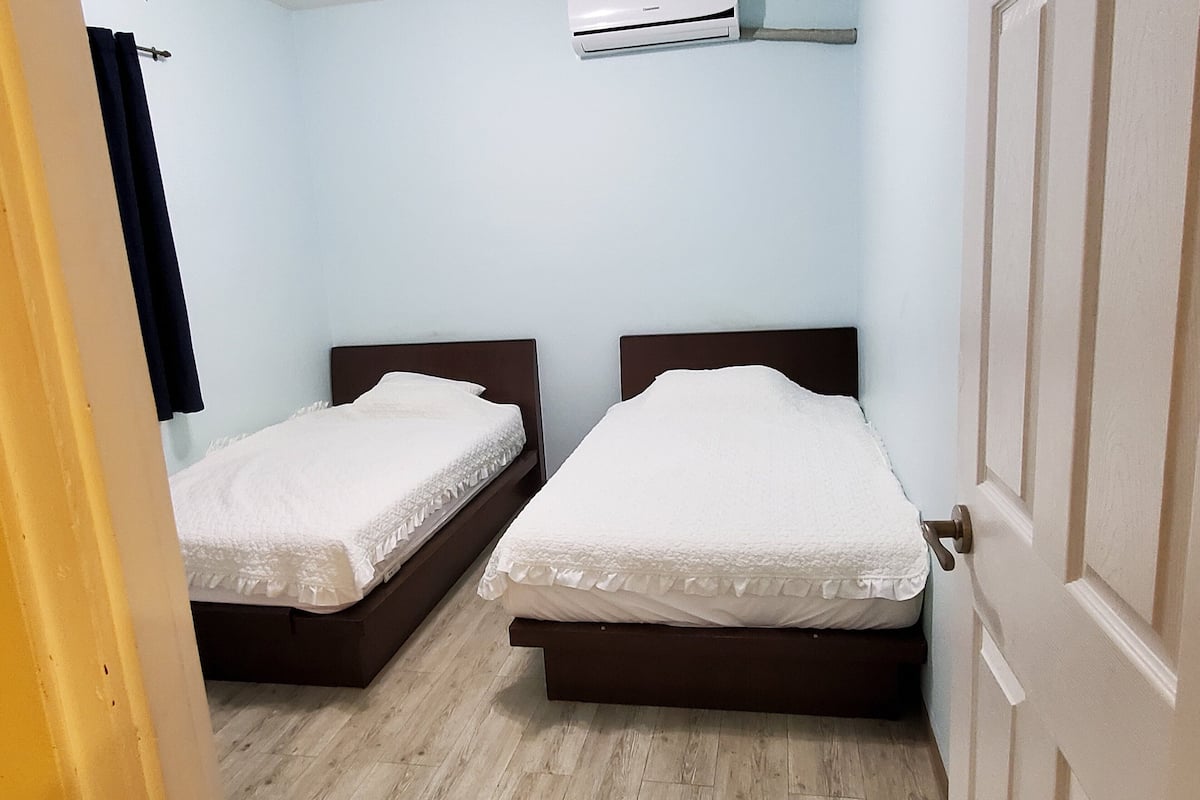 A room with two single beds, a wooden floor, a white air conditioning unit, and a door.