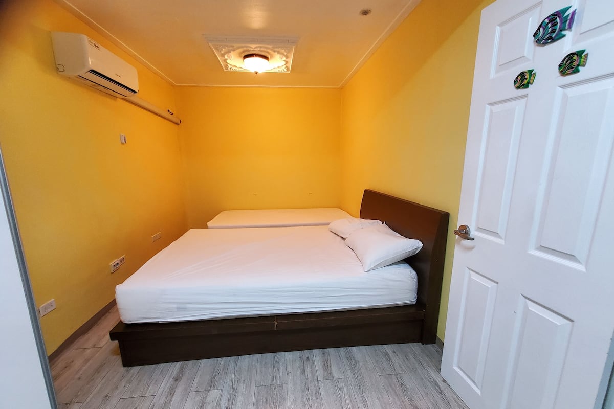 A small, single-bed room with a wooden floor, a white door, and an air conditioning unit on the wall.