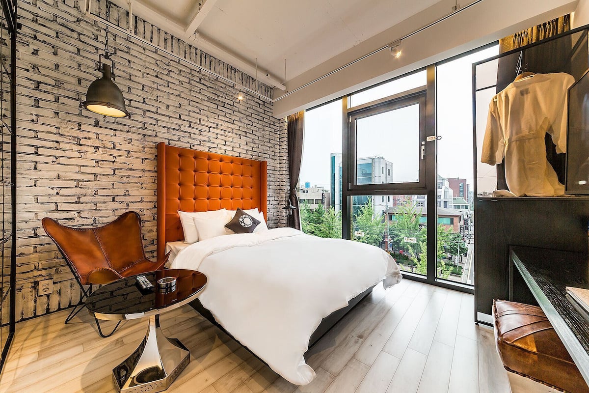 A modern hotel room with a brick-patterned wall, a tufted headboard, a large window with a city view, a glass table, and a leather chair.