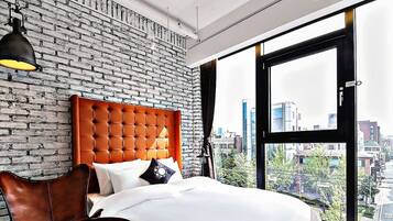 A modern hotel room with a brick wall, a large window with a city view, a tufted orange headboard, and a hanging lamp.