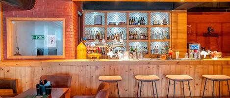 A bar area with wooden stools, a counter with bottles, and a sign indicating social distancing.