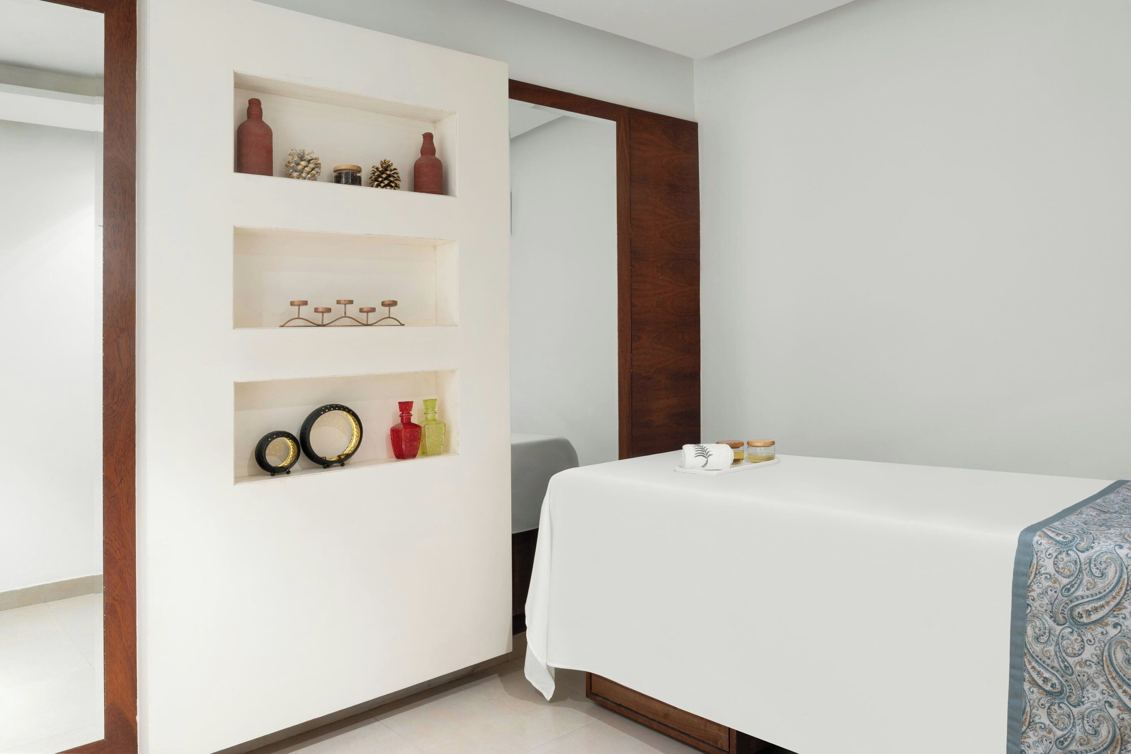 A hotel room with a bed, a white wall with shelves containing decorative items, and a wooden door frame.