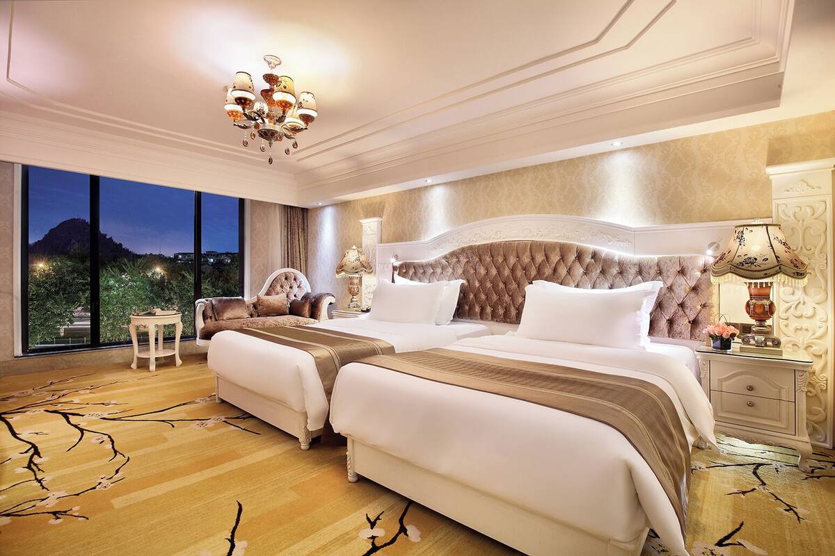 A luxurious bedroom with two beds, a chandelier, and a nightstand with a lamp.