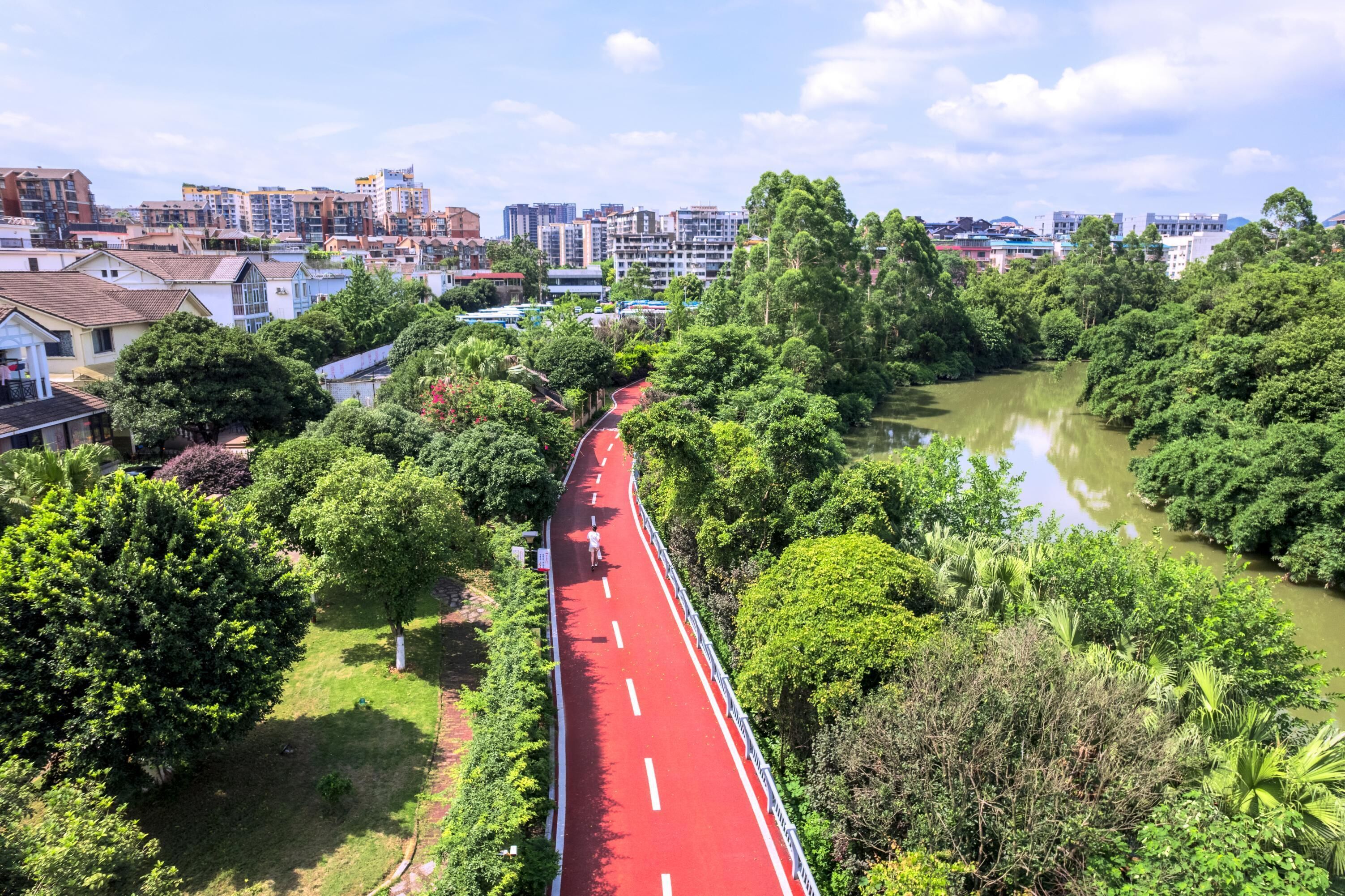 A red pathway through a lush green area with a river on one side and buildings in the background.