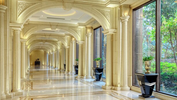 A grand hallway with marble floors, classical columns, and large windows offering a view of greenery.
