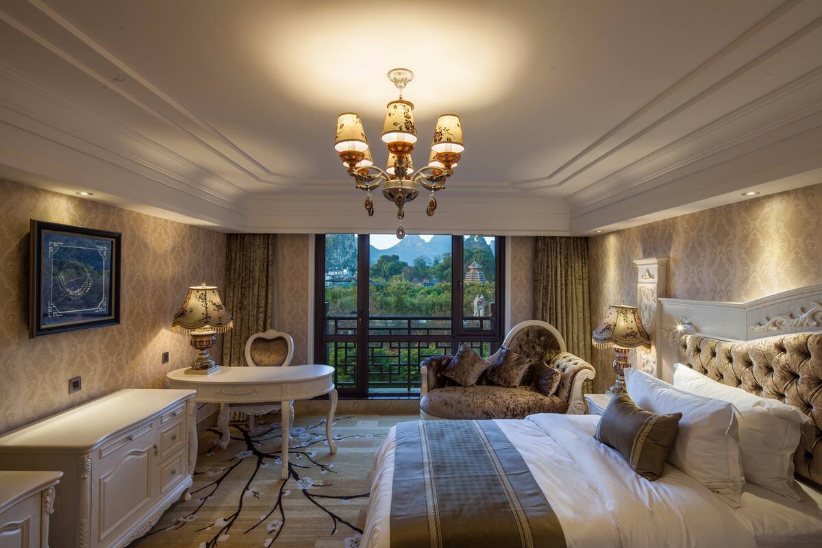 A luxurious bedroom with a large bed, a chandelier, a desk, and a view of greenery outside.
