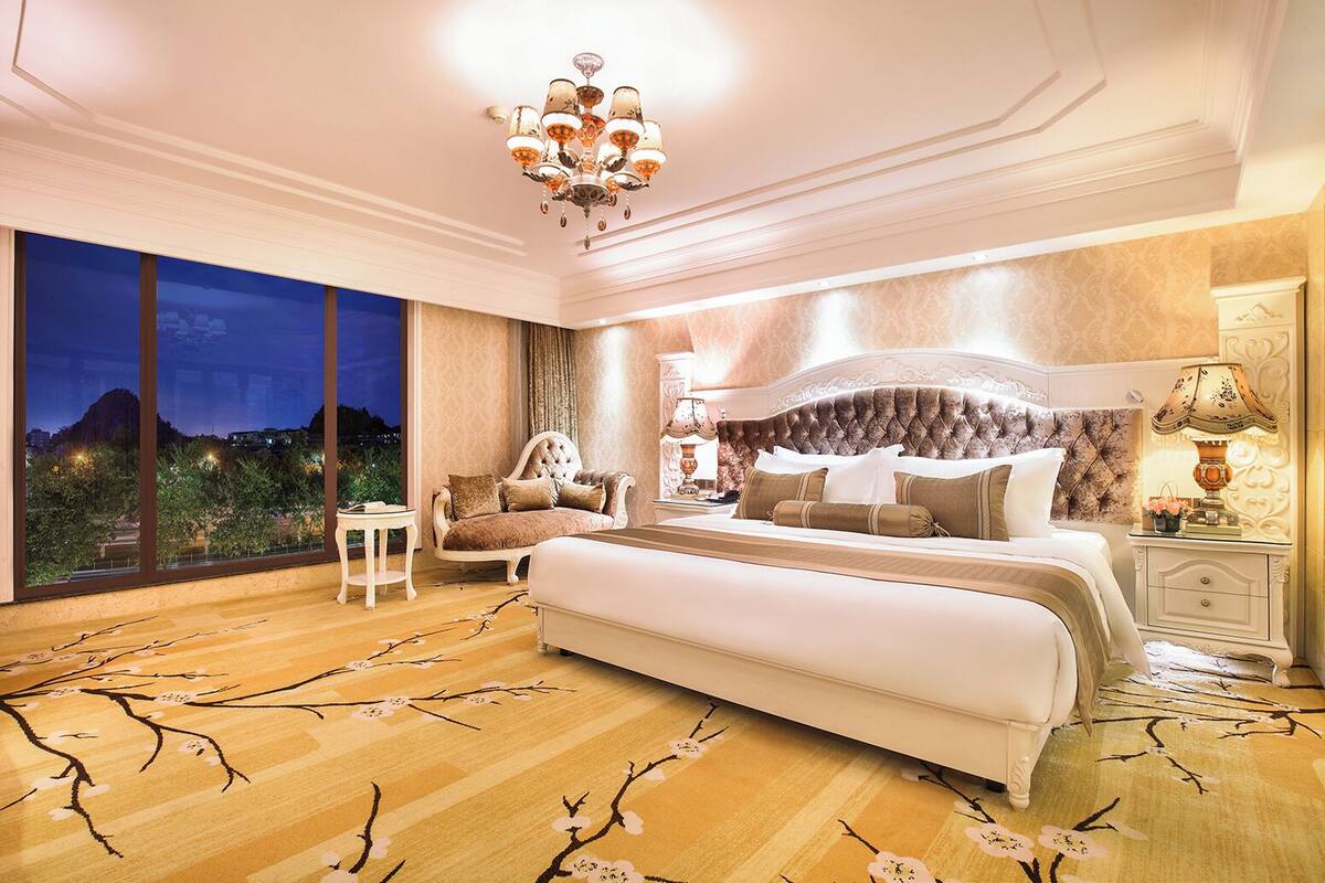 A bedroom with a large bed, a nightstand, a chair, and a view of the city at dusk.