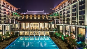 A hotel with a traditional architectural style, featuring a central courtyard with a swimming pool, multiple balconies, and a well-lit facade.
